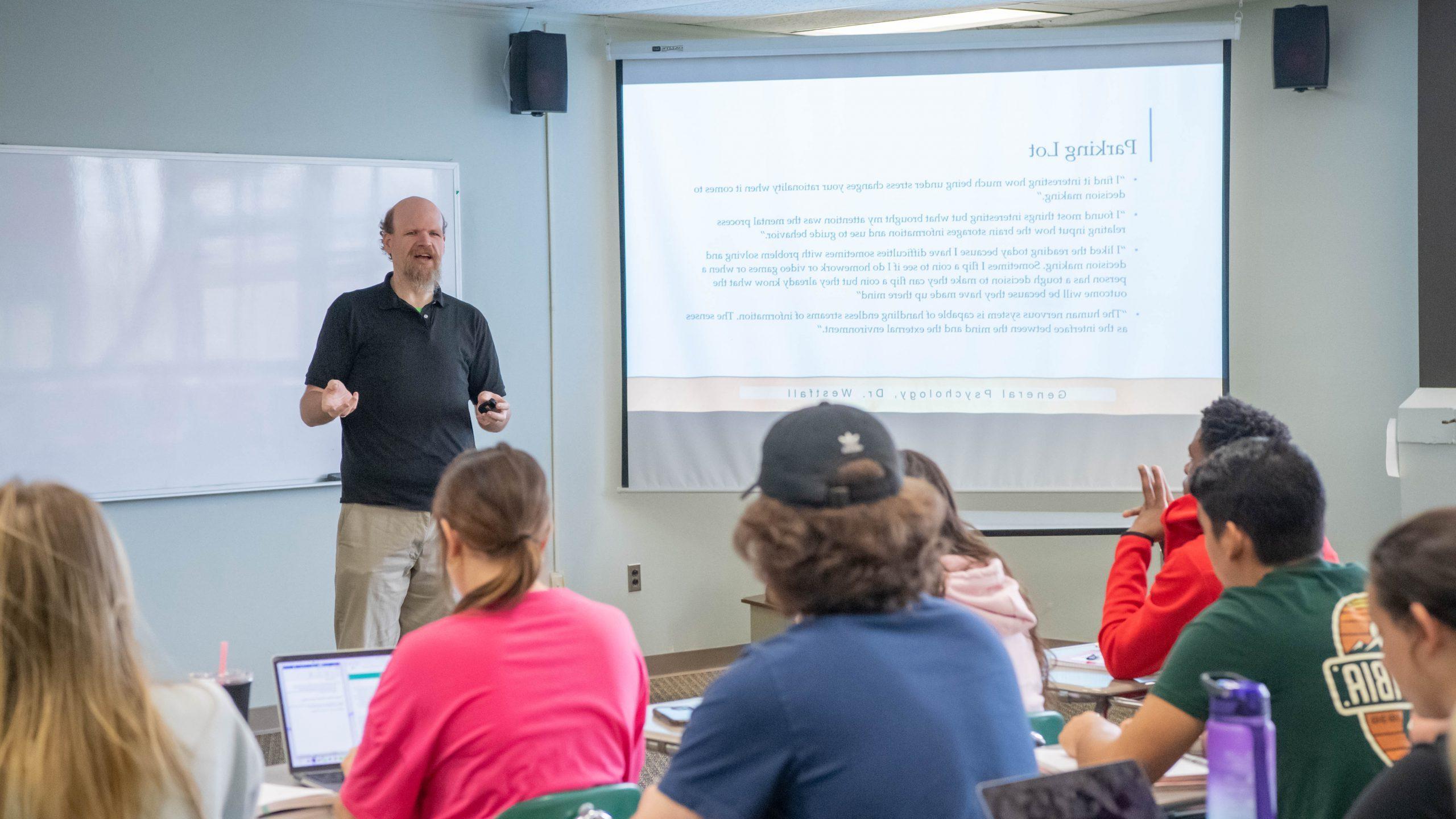 Psychology professor standing before class and teaching from a PowerPoint presentation that's displayed on a projector screen before the students.