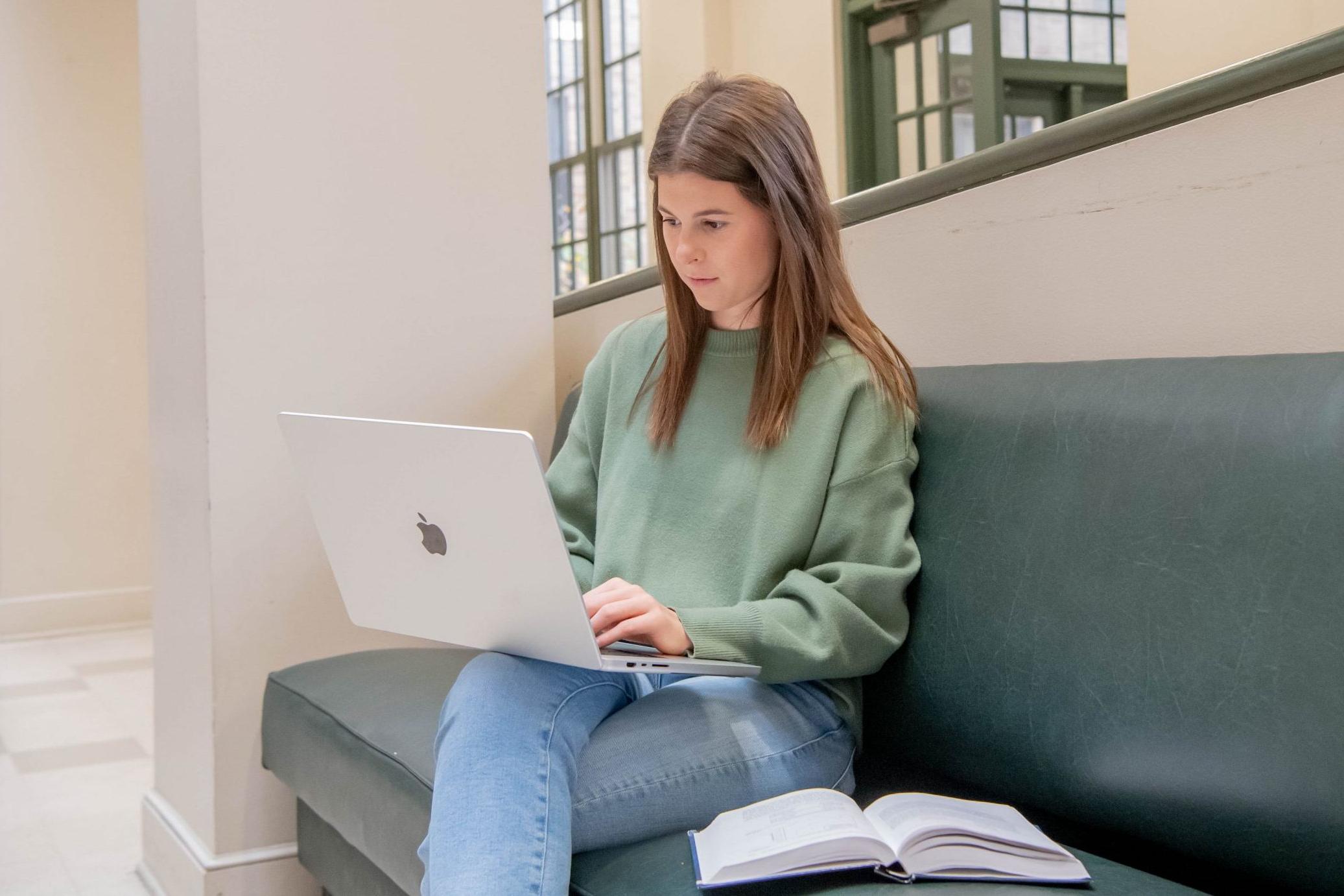 Student sitting on a bench with laptop in lap and open textbook on the bench in the College of Business lobby.