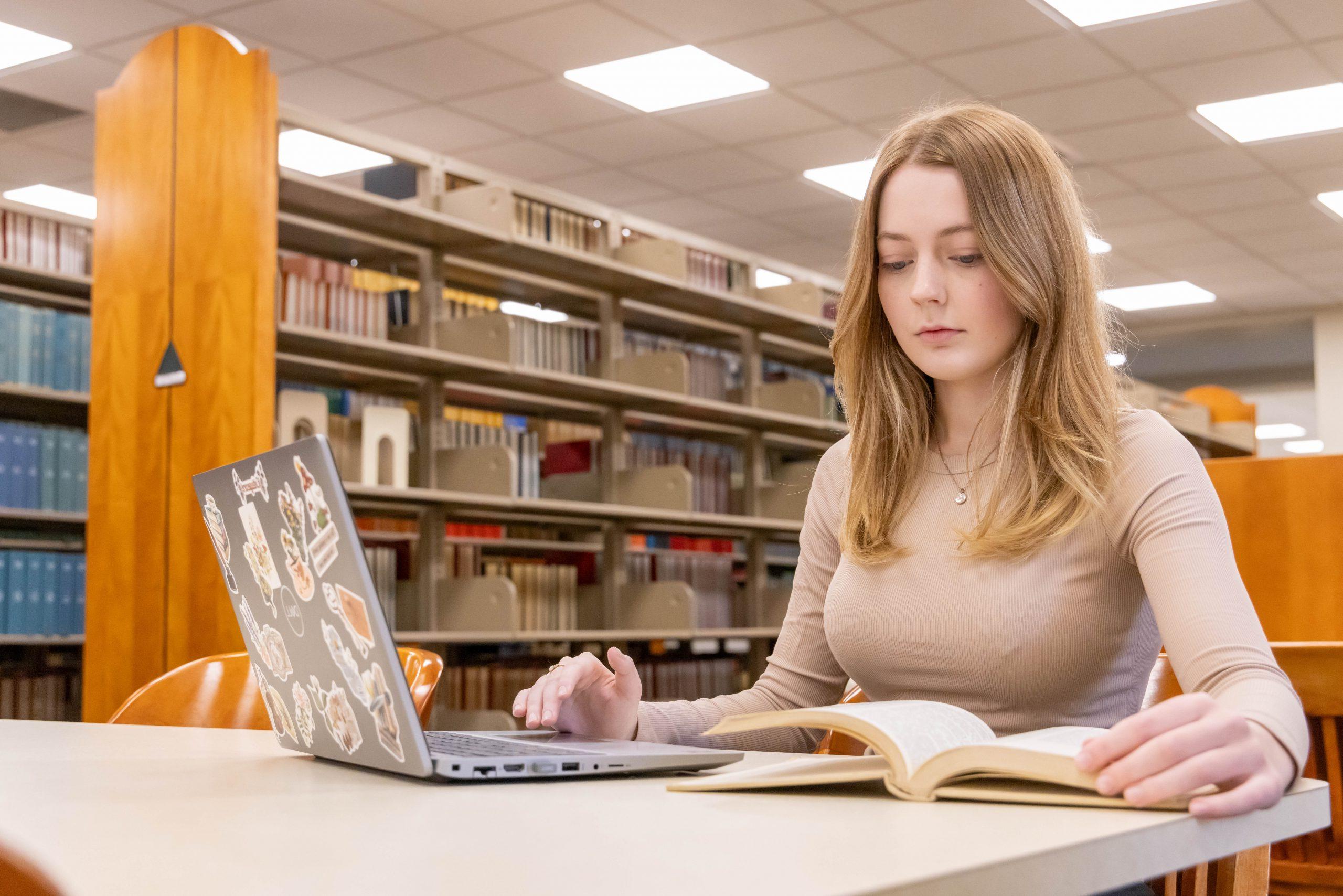 Student sitting at a table in the library while looking down at a book and laptop.